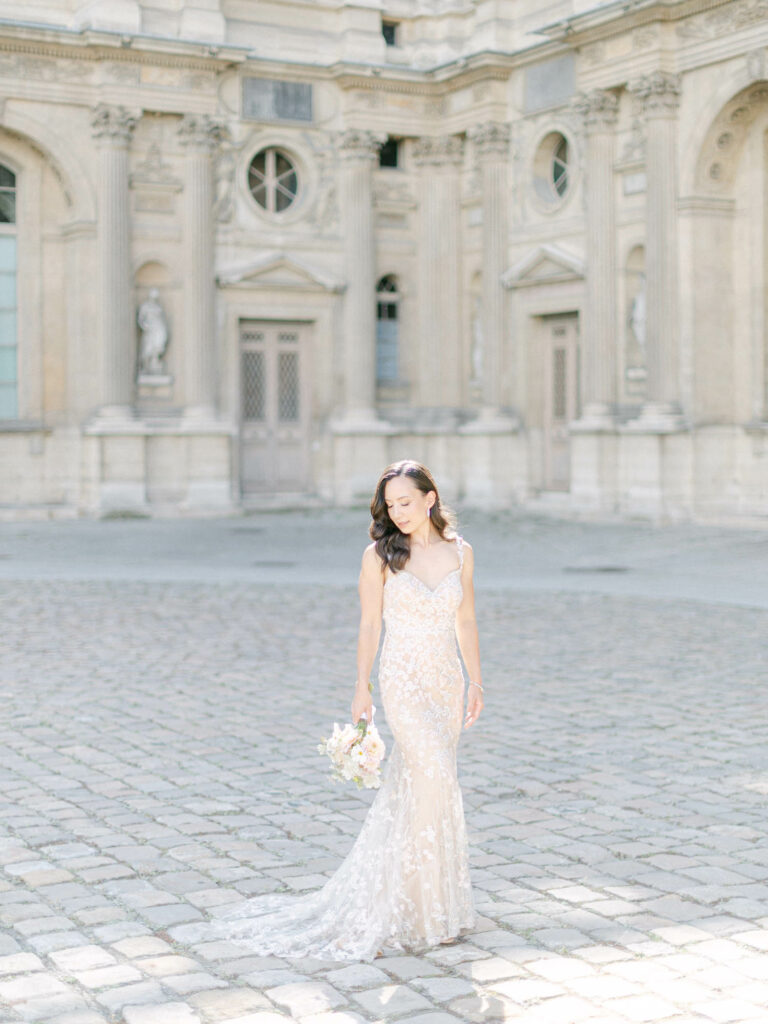 Bride in a beaded lace gown standing alone in a stone courtyard with classical architecture